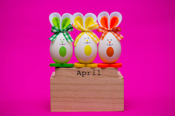 Разноцветные смешные яйца в виде милых кроликов с деревянным календарем, установленным на апрельском изолированном розовом фоне. Счастливой пасхальной открытки. Апрель дурацкий день. Праздничное оформление
 - Фото, изображение