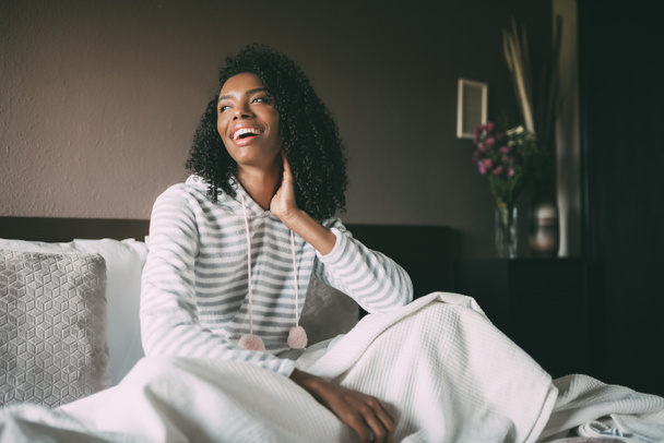 Pretty black woman covered with sheet and sit on bed looking away - Photo, image