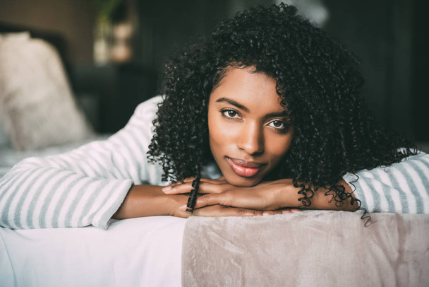 close up of a pretty black woman with curly hair smiling and lying on bed looking at the camera - Photo, Image