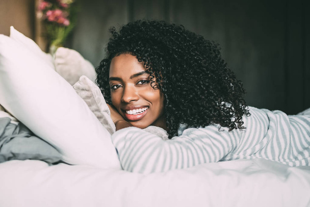 close up of a pretty black woman with curly hair smiling and lying on bed looking at the camera - Foto, Imagem