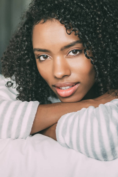 close up of a pretty black woman with curly hair smiling and lying on bed looking at the camera - Zdjęcie, obraz