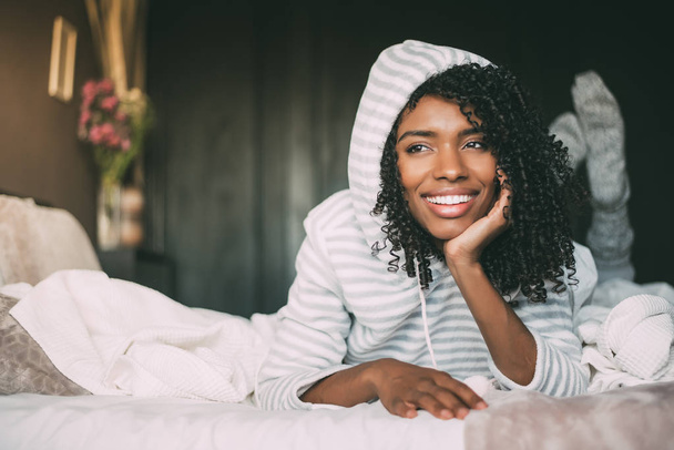 lähikuva kaunis musta nainen kihara tukka hymyillen ja makaa sängyllä katsellen pois
 - Valokuva, kuva