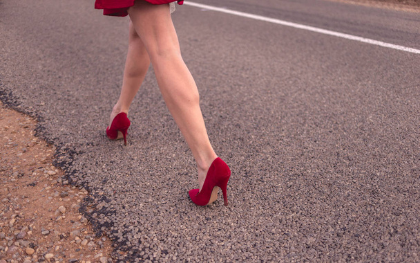 schöne Beine einer Frau, die einen kurzen Rock und rote Schuhe trägt und die Fahrbahn hinunterschreitet  - Foto, Bild