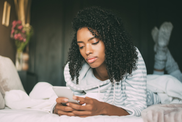 Beautiful serious thoughtful and sad black woman with curly hair using smartphone on bed - Foto, Bild