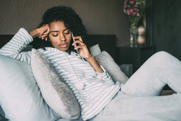 schöne ernsthafte nachdenkliche und traurige schwarze Frau mit lockigem Haar mit Smartphone auf dem Bett - Foto, Bild