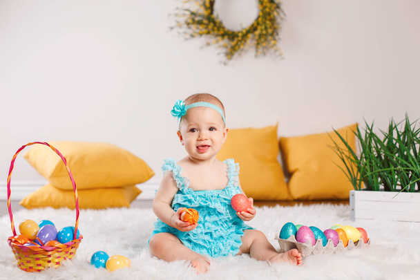 Cute adorable Caucasian baby girl in blue green romper sitting on white soft fluffy rug carpet in studio. Kid child playing with Easter colorful eggs celebrating traditional holy Christian holiday. - Foto, Imagem