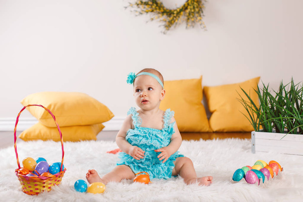 Χαριτωμένο αξιολάτρευτο μωρό Καυκάσου κορίτσι σε μπλε πράσινο ΣΑΛΟΠΕΤΑ κάθεται σε λευκή μαλακή χνουδωτή κουβέρτα ταπήτων στο studio. Παιδί παιδί παίζει με πολύχρωμα αυγά του Πάσχα γιορτάζουμε παραδοσιακά ιερό χριστιανική γιορτή. - Φωτογραφία, εικόνα
