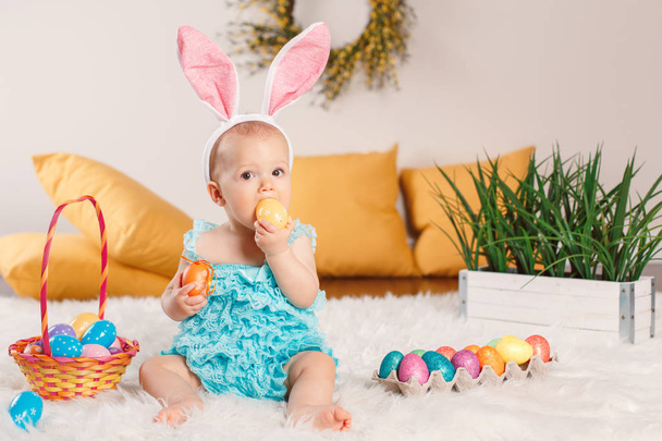 Cute adorable Caucasian baby girl in blue green romper wearing bunny ears sitting in studio. Kid child playing with Easter colorful eggs celebrating traditional holy Christian holiday. - Photo, Image