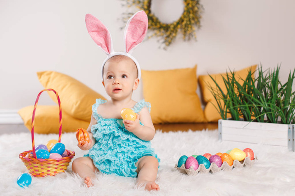 Cute adorable Caucasian baby girl in blue green romper wearing bunny ears sitting in studio. Kid child playing with Easter colorful eggs celebrating traditional holy Christian holiday. - Photo, Image