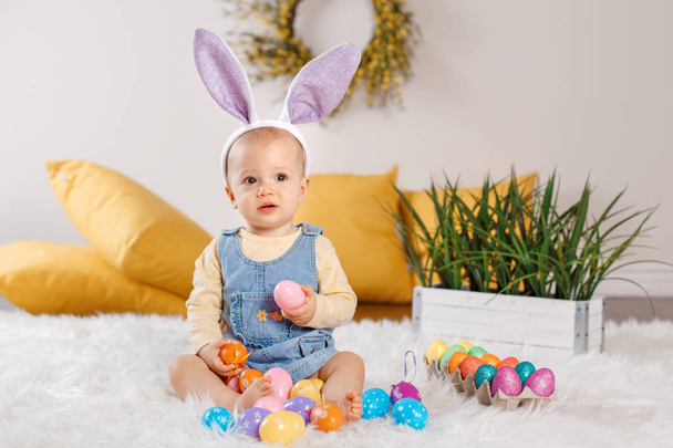 Cute adorable Caucasian baby girl in jeans dress wearing purple bunny ears sitting in studio. Kid child playing with Easter colorful eggs celebrating traditional holy Christian holiday. - Photo, Image