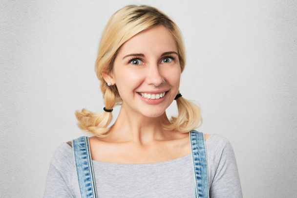 В приміщенні знімок молодої блондинки з проклятим носом, носить комбінезон, просто зачіска посміхається і показує свої зуби всьому світу. Ізольовані над білою бетонною стіною
 - Фото, зображення