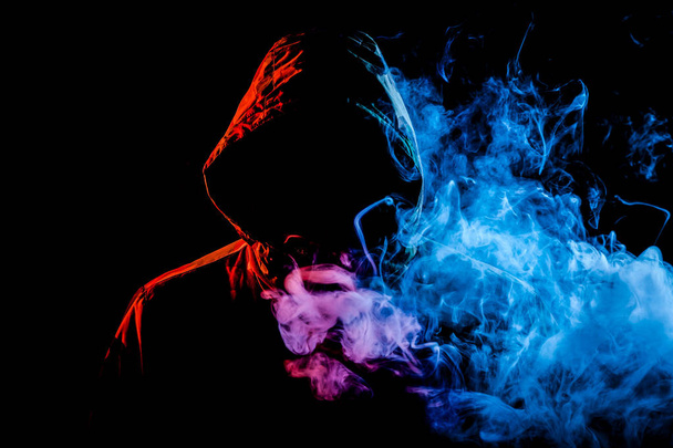 Mladý muž v černé kápi kouří a dýchá z barevné zelená a růžová kouře z vape na černém pozadí izolované - Fotografie, Obrázek