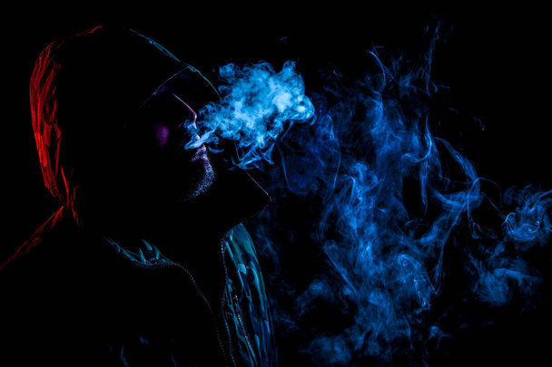 Ένας νεαρός άνδρας σε μια μαύρη κουκούλα καπνίζει ένα vape και αναδίδει ένα χρωματιστό μπλε καπνό πάνω σε μαύρο φόντο απομονωμένες - Φωτογραφία, εικόνα