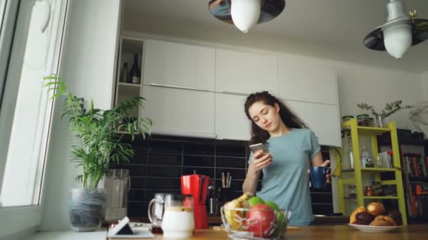 Attractive happy girl dancing in kitchen while browsing social media on smartphone and listening to music at home in the morning - 映像、動画