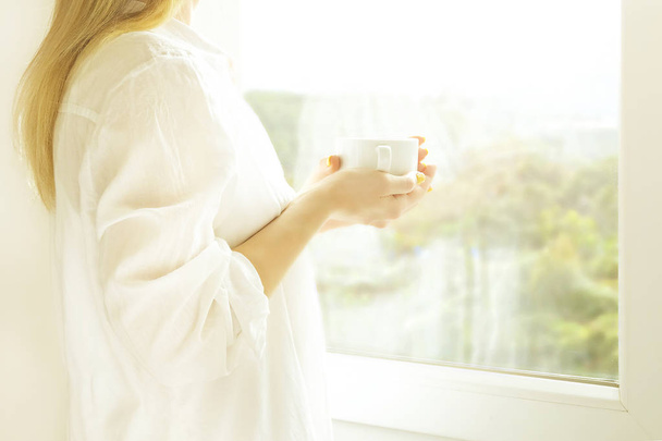 Big spacey hotel room full of sunlight and sun beams. Optimistic start of the day. Blond woman cozy home clothing welcoming the morning daylight. New day new me concept. - Photo, Image