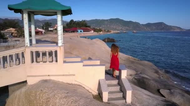 wonderful aerial view blonde girl in red dress waved by wind walks along stone bridge above ocean - Кадри, відео