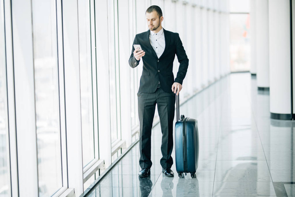 Элегантный бизнесмен проверяет электронную почту на мобильном телефоне во время прогулки с чемоданом внутри терминала аэропорта. Опытный мужчина работодатель с помощью мобильного телефона перед поездкой на работу
 - Фото, изображение