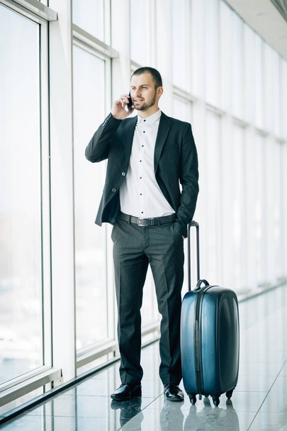 Täyspitkä muotokuva onnellinen mies kävely matkalaukku ja puhelin terminaalin lentokentällä
 - Valokuva, kuva