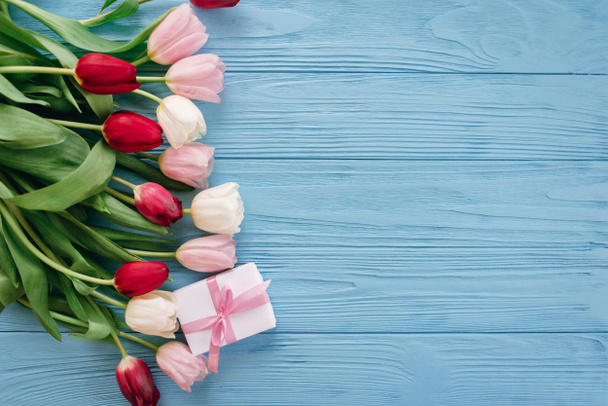 Floral composition of colourful tulips laying in row on left side on blue wooden background with gift box - Photo, Image