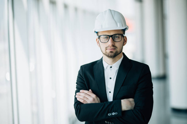 erfolgreicher Ingenieur oder Architekt, fröhlicher Geschäftsmann mit verschränkten Armen, der einen Helm auf dem Hintergrund eines Bürogebäudes trägt - Foto, Bild
