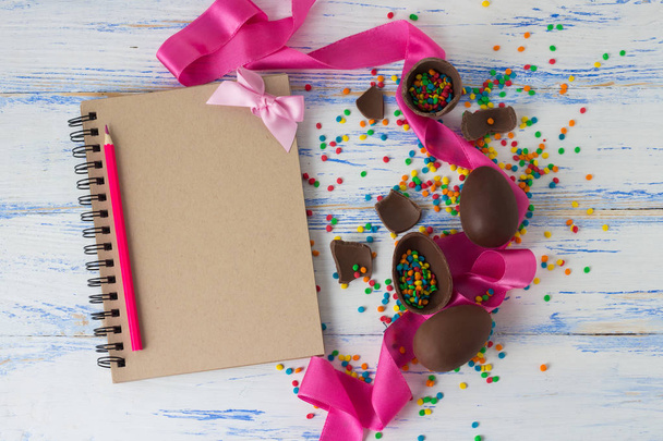 Πασχαλινά σοκολατένια αβγά, ροζ κορδέλα, το σημειωματάριο και χρωματιστά μολύβια, - Φωτογραφία, εικόνα