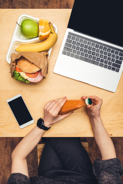 cropped image of woman opening bottle of juice at table with food and gadgets - Foto, afbeelding