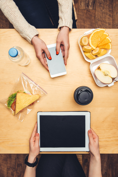 cropped image of women sitting at table with take away food and using gadgets  - Foto, afbeelding