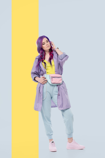 創造的な背景の上にヘッドフォンで聴く音楽を紫のトレンチ コートでスタイリッシュな若い女性 - 写真・画像