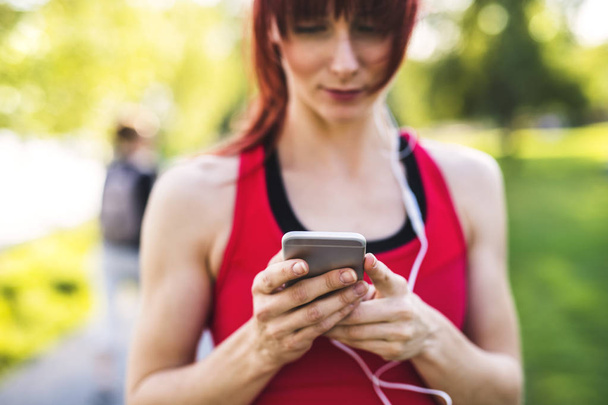 Young athlete in park holding smart phone, listening to music. - Φωτογραφία, εικόνα