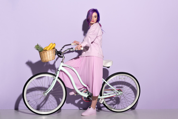 Zijaanzicht van de jonge vrouw in roze kleding op fiets met ananas en bananen in mand - Foto, afbeelding