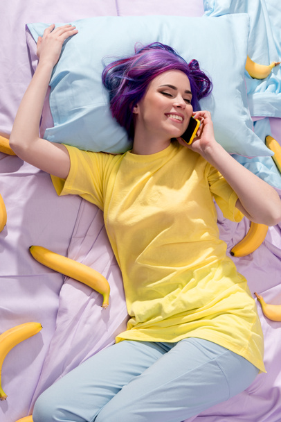 high angle view of happy young woman lying in bed with bananas and talking by phone - Фото, изображение