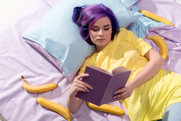 high angle view of young woman lying in bed with bananas and reading book - Photo, Image