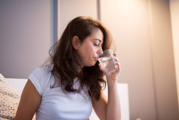 病気の女性、自宅のベッドに座って水を飲むと頭痛に苦しんで  - 写真・画像