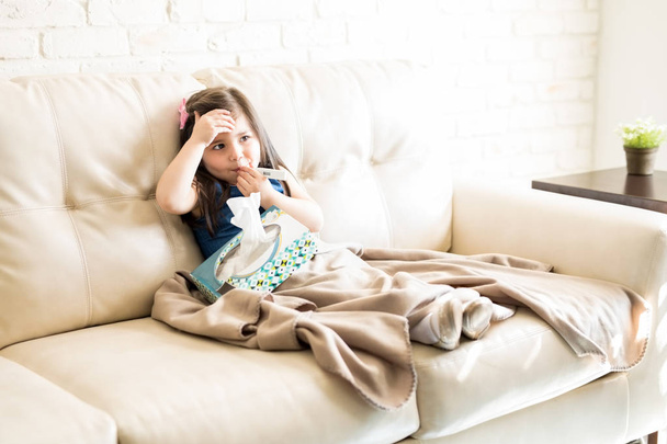 little girl holding thermometer in mouth and holding box of tissues on sofa in living room - Photo, Image