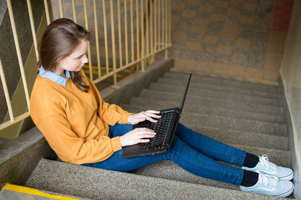 Young female college student sitting on stairs at school, writing essay on her laptop. Education concept. - Foto, Bild