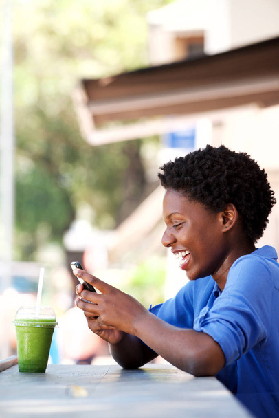携帯電話のテキスト メッセージを読むカフェに座って幸せな若い黒人女性の側面の肖像画  - 写真・画像