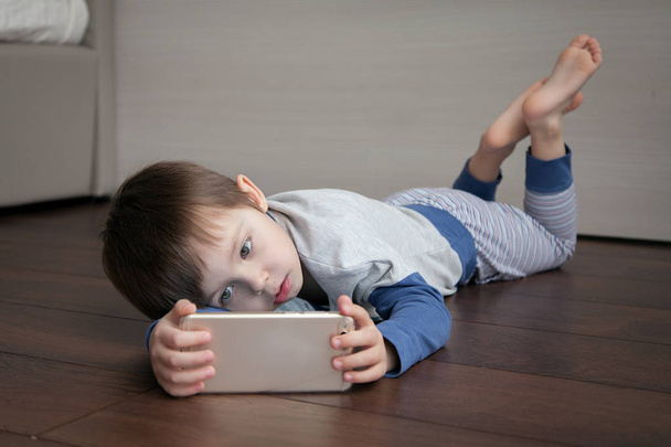 A child lying on the floor looks at something interesting in the gadget.  - Φωτογραφία, εικόνα