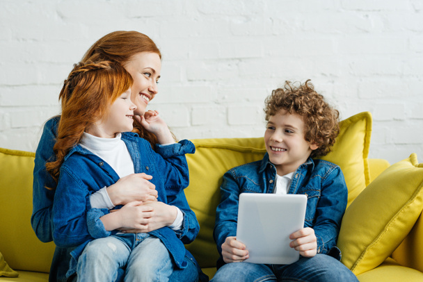 Hugging mother and daughter sitting on sofa with son holding tablet - Photo, Image