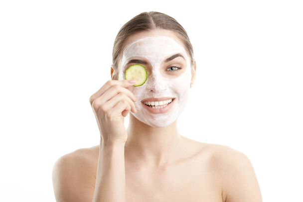 Close-up shot of a smiling beautiful young woman wearing a face mask and holding a cucumber slice in front of her eye. Isolated on white background.  - Фото, изображение