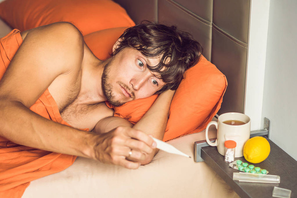 Young man feeling cold, lying in bed, drinking tea and measuring temperature with thermometer - Foto, Bild