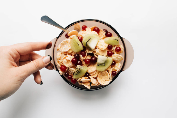 female hand holding bowl with delicious crispy cornflakes, kiwi pieces and cranberries on white background, closeup, healthy breakfast  - Photo, Image