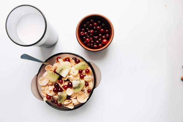 köstliche knusprige Cornflakes mit Kiwi-Stücken und Preiselbeeren, Glas Milch auf weißem Hintergrund, Nahaufnahme, gesundes Frühstück  - Foto, Bild