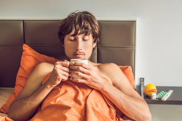 Young man feeling cold, lying in bed, drinking tea and measuring temperature with thermometer - Photo, Image
