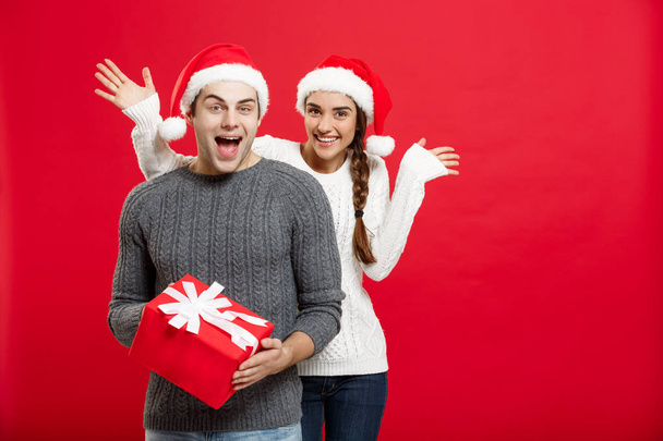 Concepto de Navidad - Mujer joven cubriendo los ojos de los hombres con la mano y dando gran regalo sorpresa. Aislado sobre fondo rojo
 - Foto, imagen