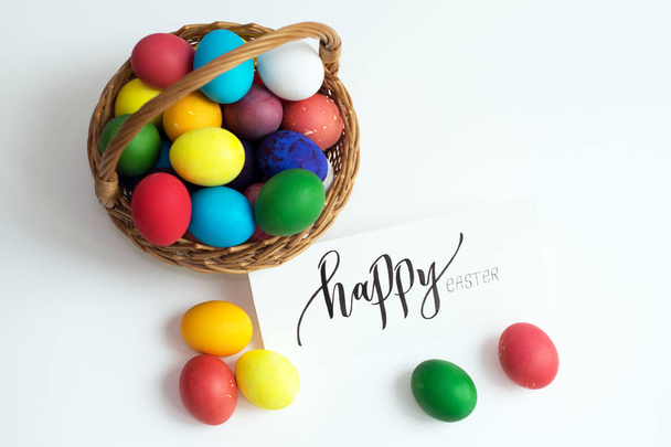 Easter card with colorful Easter eggs in a basket and calligraphic inscription "Happy Easter", bright colored eggs on a white table - Photo, Image
