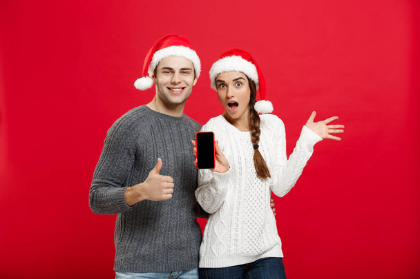 Vánoční koncept - šťastný mladý pár v vánoční svetry ukazující bušit se gesto s mobilním telefonem - Fotografie, Obrázek