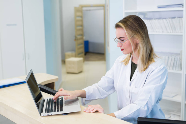 Улыбающаяся женщина-врач, использующая компьютер в своем кабинете в больнице. Молодая привлекательная кавказка врач-терапевт в медицинской форме сидит за столом
 - Фото, изображение