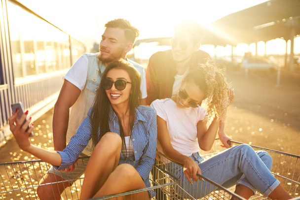Selfie time! Group of happy young people having fun on shopping trolleys. Multiethnic young people racing on shopping cart. Beautiful summer day with sunlight. Lifestyle concept  - Foto, imagen