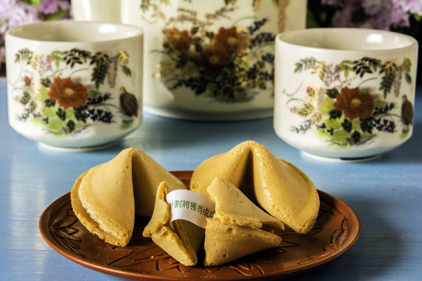 um prato oriental vermelho de três biscoitos da sorte chineses em quebrado expondo fortuna de papel e duas xícaras de chá e panela de chá na mesa de madeira azul com fundo de flor
 - Foto, Imagem