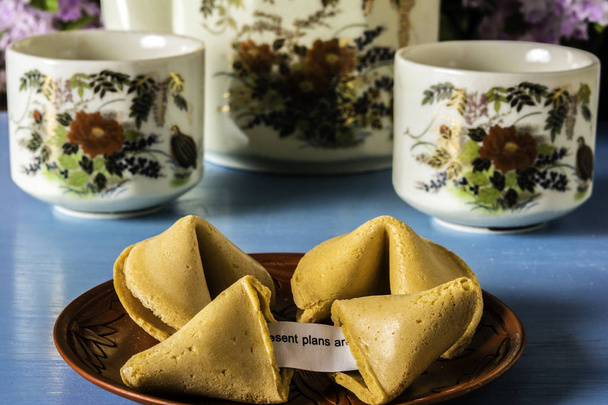 un piatto orientale rosso di tre biscotti cinesi fortuna su rotto esponendo fortuna carta e due tazze da tè e teiera su tavolo di legno blu con sfondo fiore
 - Foto, immagini